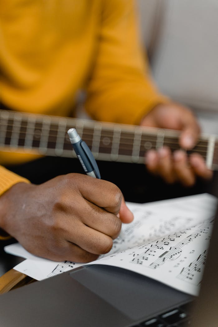 Close-up of a person writing on sheet music while holding a guitar, focusing on creativity and musical composition.