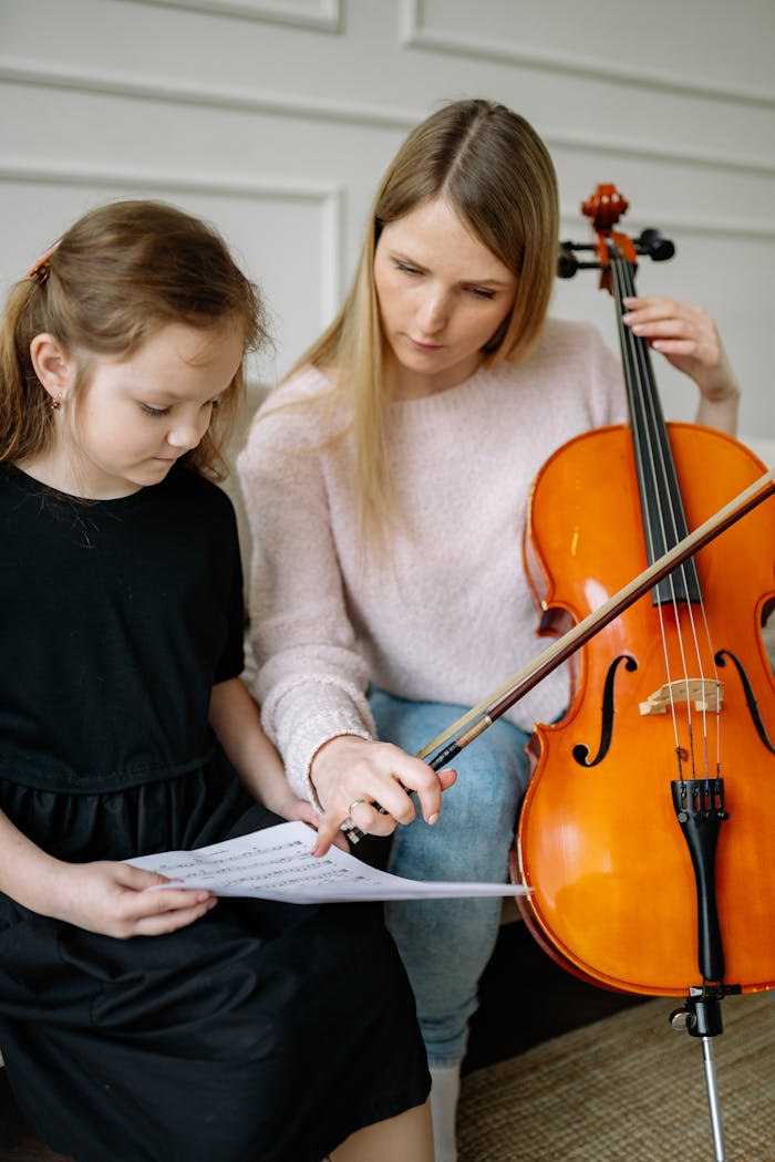 Woman teaching young girl to play cello at home, focusing on music sheet.